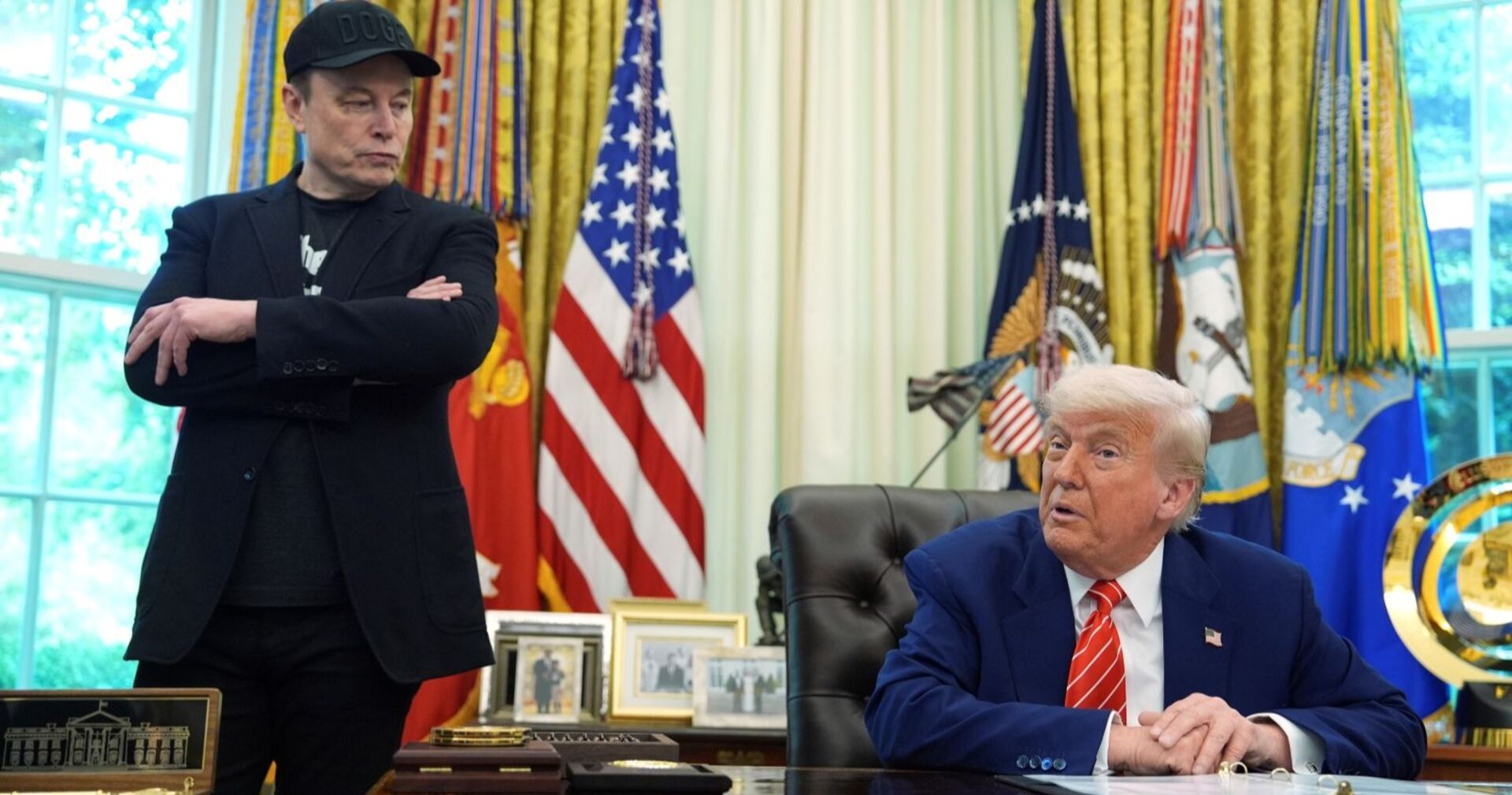 Elon Musk steht mit verschränkten Armen neben Donald Trump, der am Schreibtisch im Oval Office sitzt. Im Hintergrund US-Flaggen und militärische Standarten.