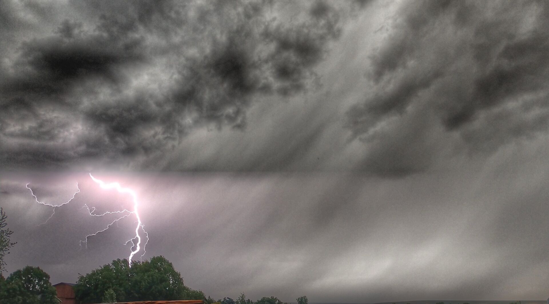 Ein heller Blitz schlägt während eines schweren Gewitters neben Bäumen ein. Der Himmel ist von dunklen, dramatischen Wolken bedeckt.