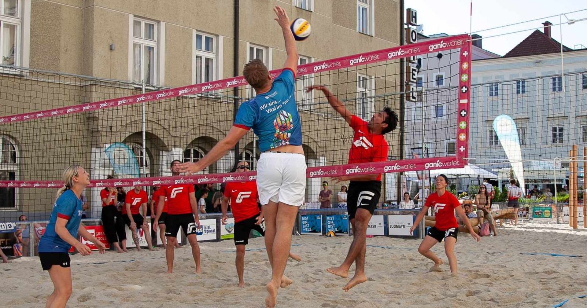 Zwei gemischte Teams spielen Beachvolleyball in einer Stadt; zwei Spieler springen am Netz um den Ball.          ChatGPT fragen