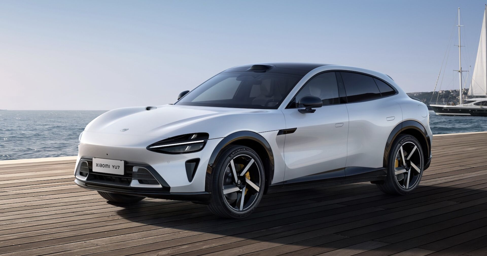 Der weiße Xiaomi YU7 Elektro-SUV steht auf einem Holzsteg vor dem Meer, im Hintergrund ein Segelboot – sportlich-elegantes Fahrzeug mit aerodynamischer Linienführung.