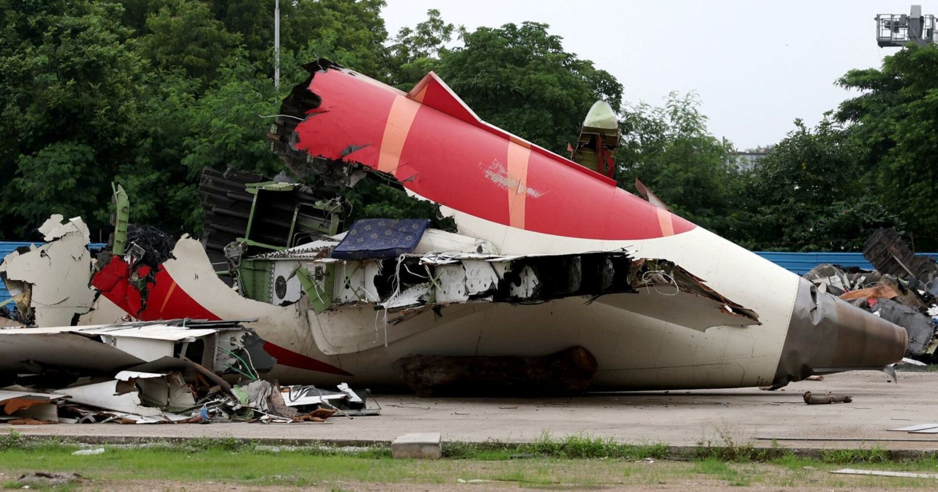 Wrackteil des Flugzeugs von Air India, stark beschädigtes Heckstück mit aufgerissenen Blechen und herausragenden Kabeln, liegt auf einem Betonplatz, im Hintergrund grüne Bäume.