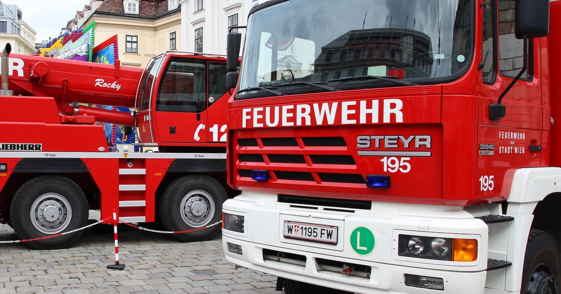 Rote Feuerwehrfahrzeuge der Stadt Wien (Steyr und Liebherr) stehen auf einem gepflasterten Platz, im Hintergrund historische Gebäude und ein buntes Fahrgeschäft.