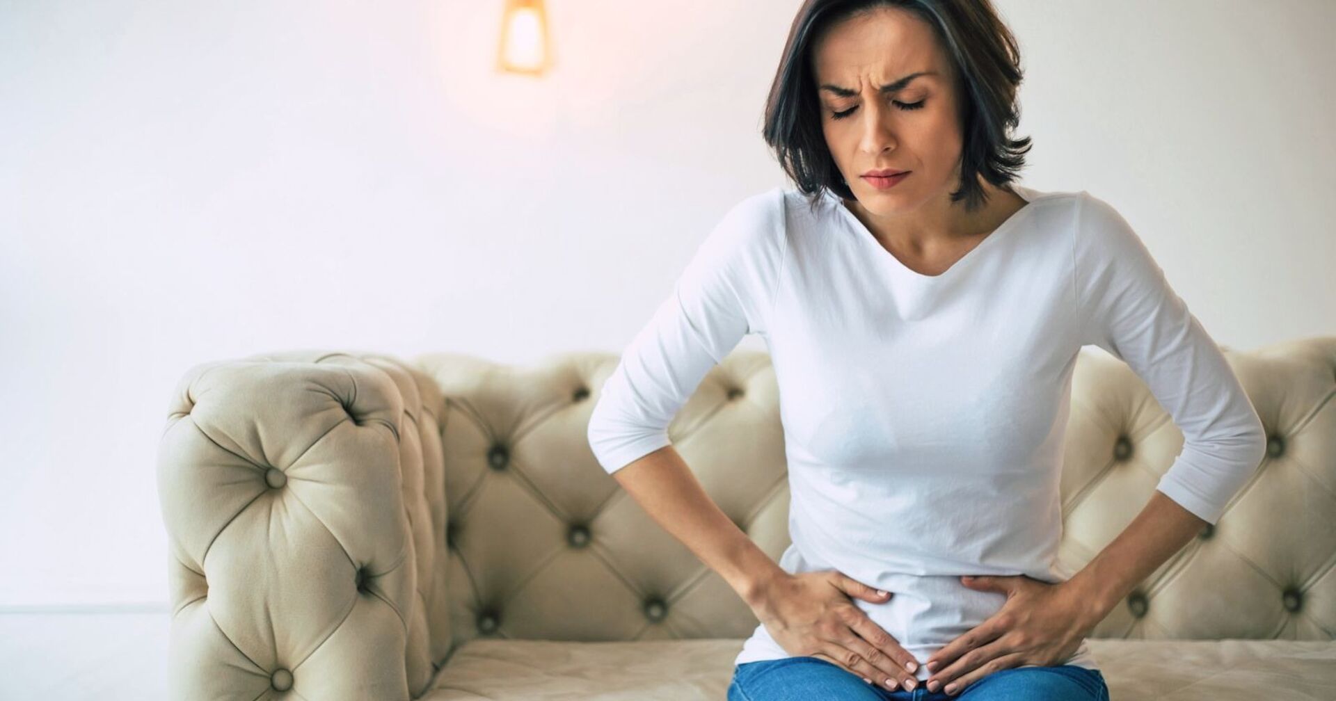 Eine Frau - bekleidet mit einem weißen, dünnen Pullover und einer Jean - sitzt auf einer Couch. Ihr Gesicht ist schmerzverzehrt und sie presst beide Hände auf ihren Unterbauch.