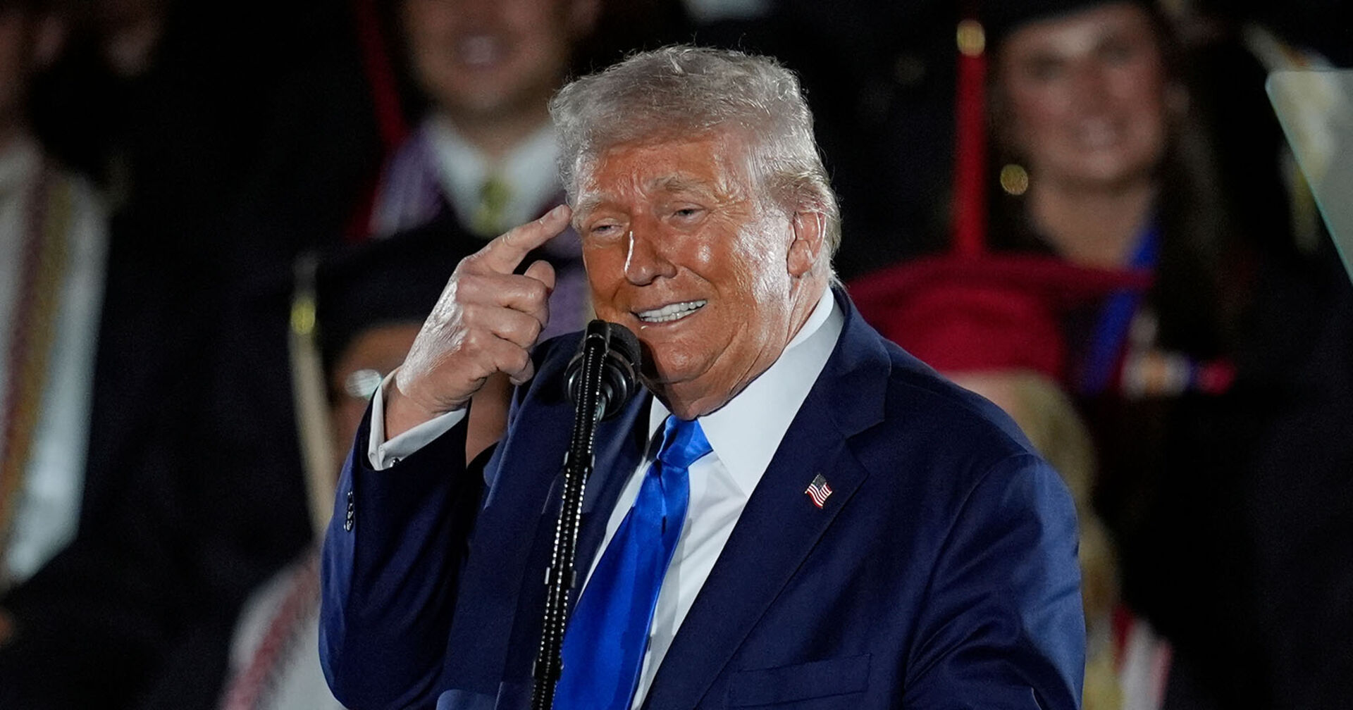 Donald Trump, im Anzug und mit Krawatte, hält sich Finger an den Kopf und zeigt "den Vogel"