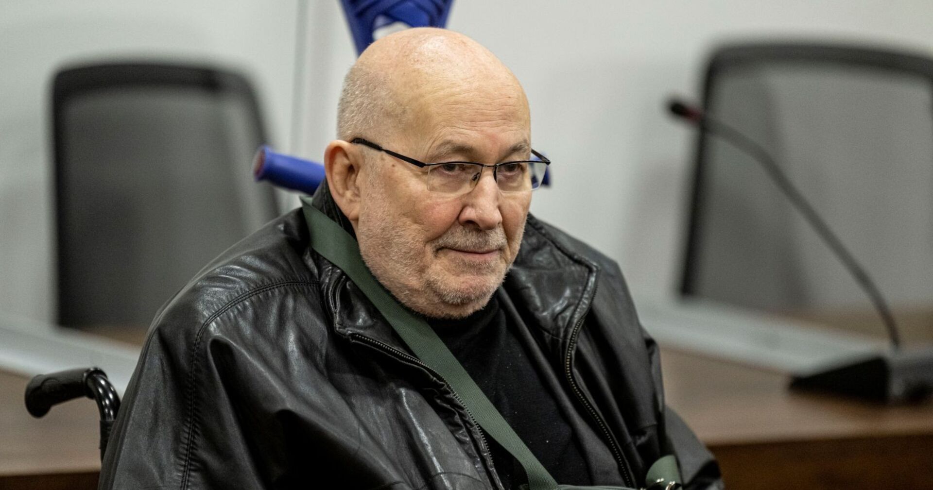 Horst Mahler sitzt im Rollstuhl, trägt eine schwarze Lederjacke und blickt ernst. Aufnahme aus einem Gerichtssaal im Jahr 2022.