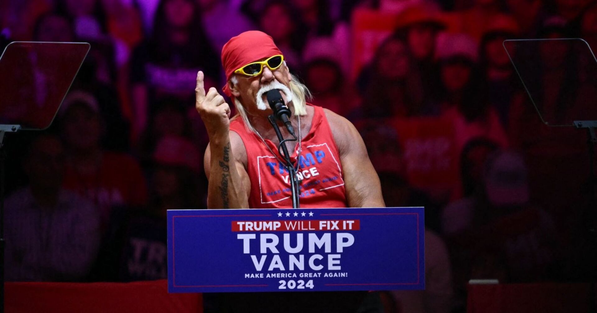 Hulk Hogan steht mit rotem Bandana und Sonnenbrille hinter einem Rednerpult mit der Aufschrift „Trump – Vance 2024“ und spricht bei einer Wahlkampfveranstaltung.
