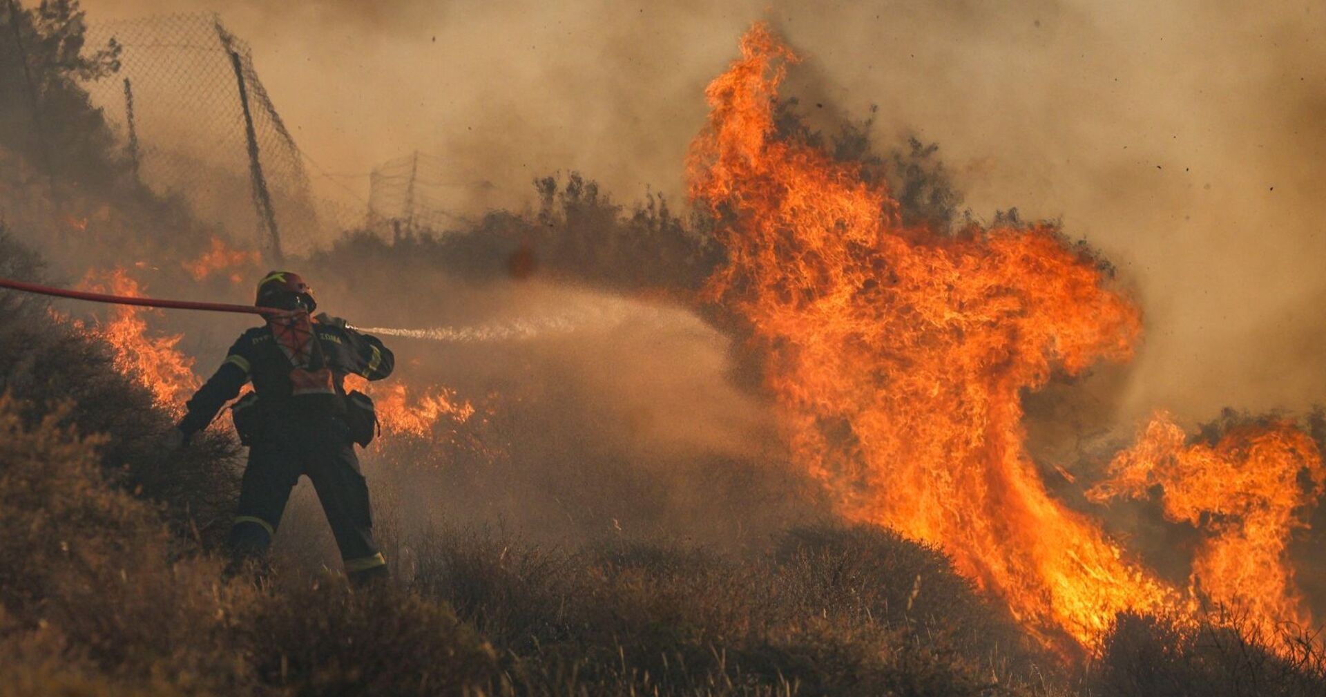 Ein Feuerwehrmann kämpft in Griechenland mit einem Wasserschlauch gegen meterhohe Flammen – dichter Rauch und Hitze erschweren den Einsatz massiv.