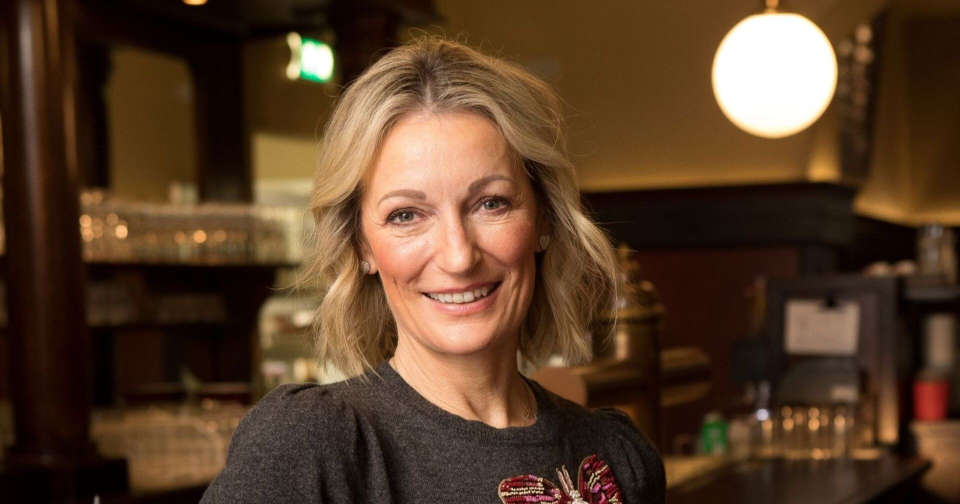 Monika Gruber lächelt in die Kamera, während sie in einer Bar steht.