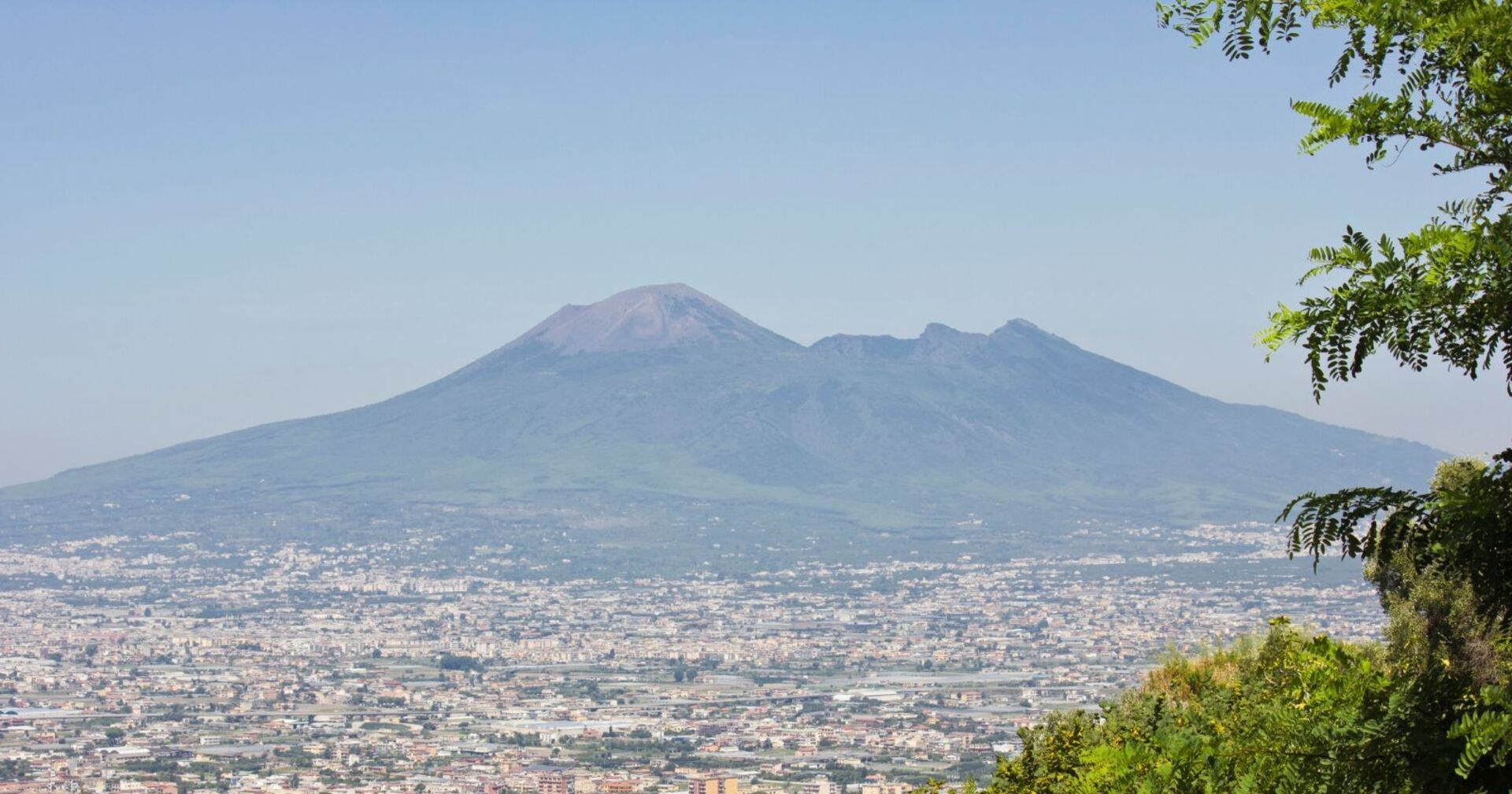 Panoramaaufnahme des Vesuvs bei Neapel, Italien, mit Blick auf die darunterliegende Stadt und grüne Vegetation im Vordergrund.