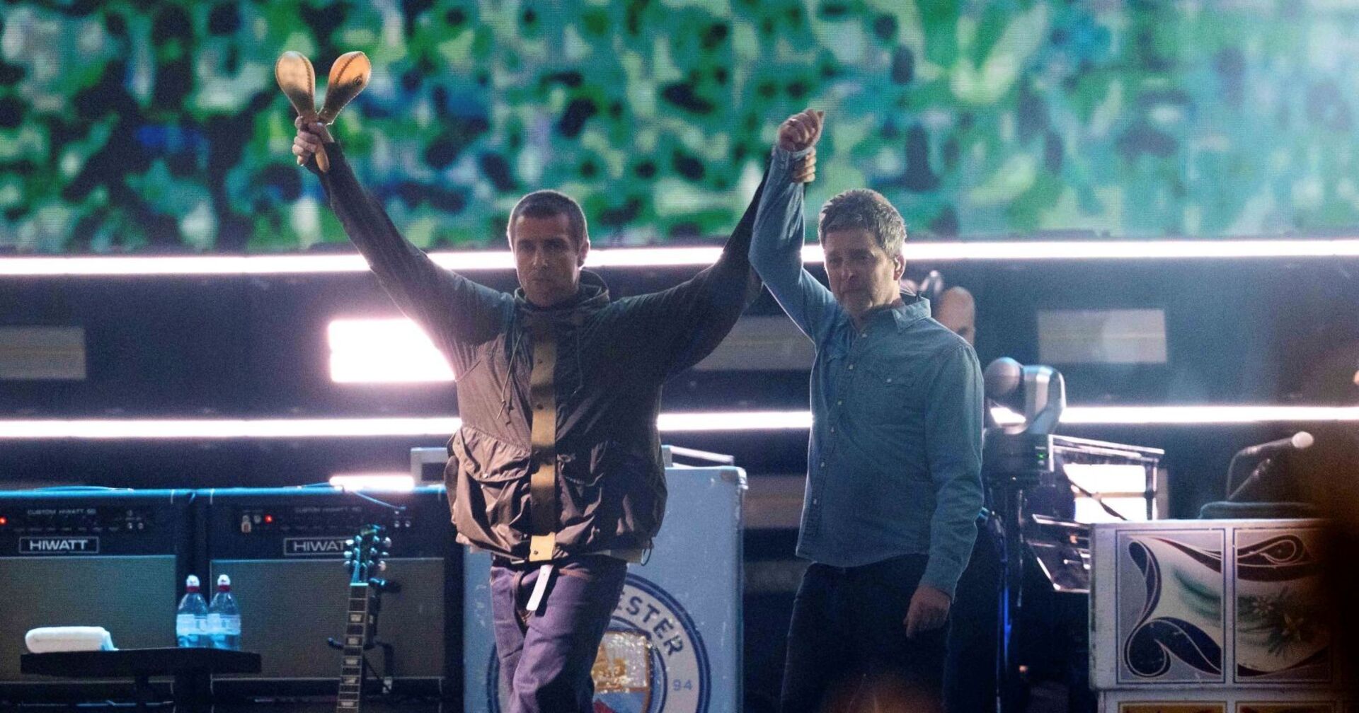Liam hält Noel Gallaghers Hand, im Hintergrund sieht man ein Wappen des Fußballclubs Manchester City
