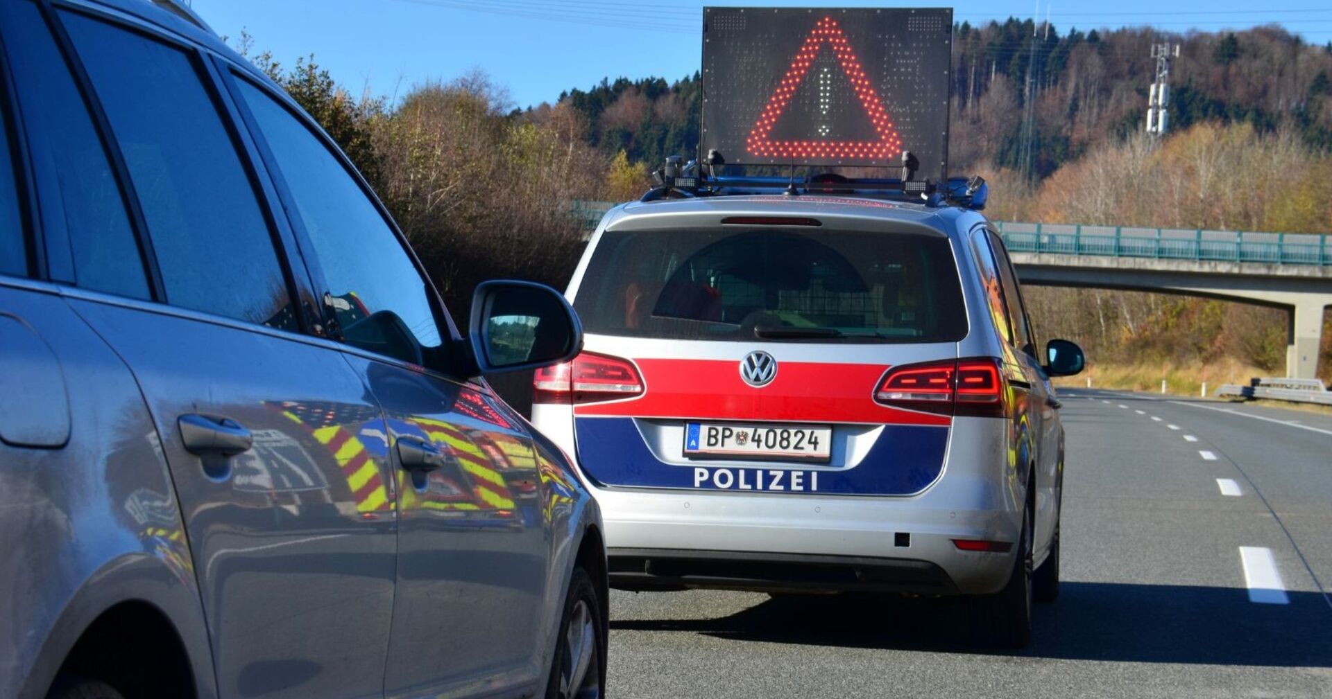 Polizeifahrzeug mit Warnanzeige und ein weiteres Auto stehen auf einer abgesperrten Landstraße in Österreich, im Hintergrund eine Brücke und Bäume.