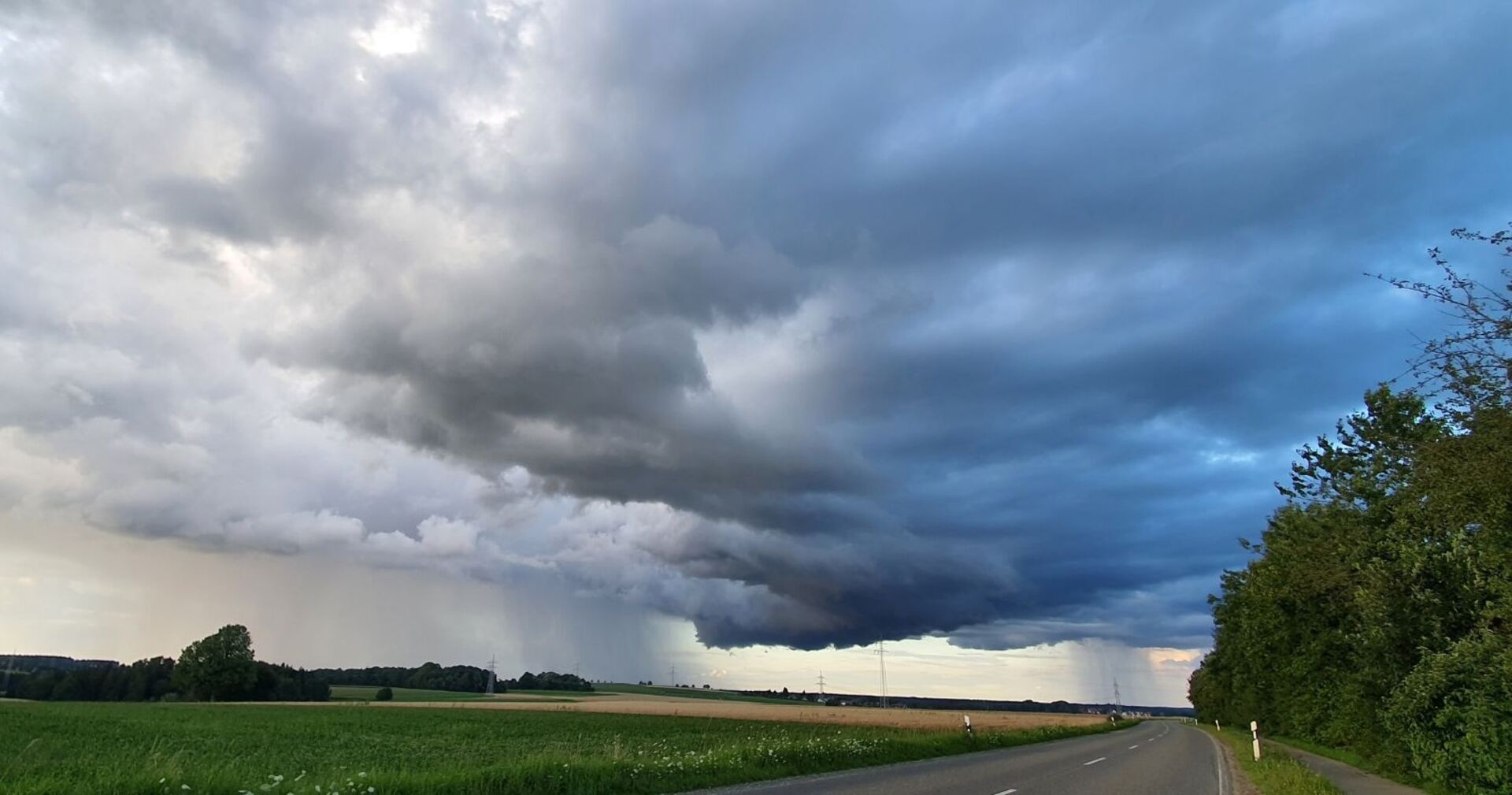 Dunkle, tief hängende Gewitterwolken über einer Landstraße und grünen Feldern, im Hintergrund ist Regen sichtbar, der aus den Wolken fällt.Bedrohliche Wolkenfront zieht über das Land – Österreich muss sich erneut auf Gewitter, Starkregen und kühle Temperaturen einstellen.