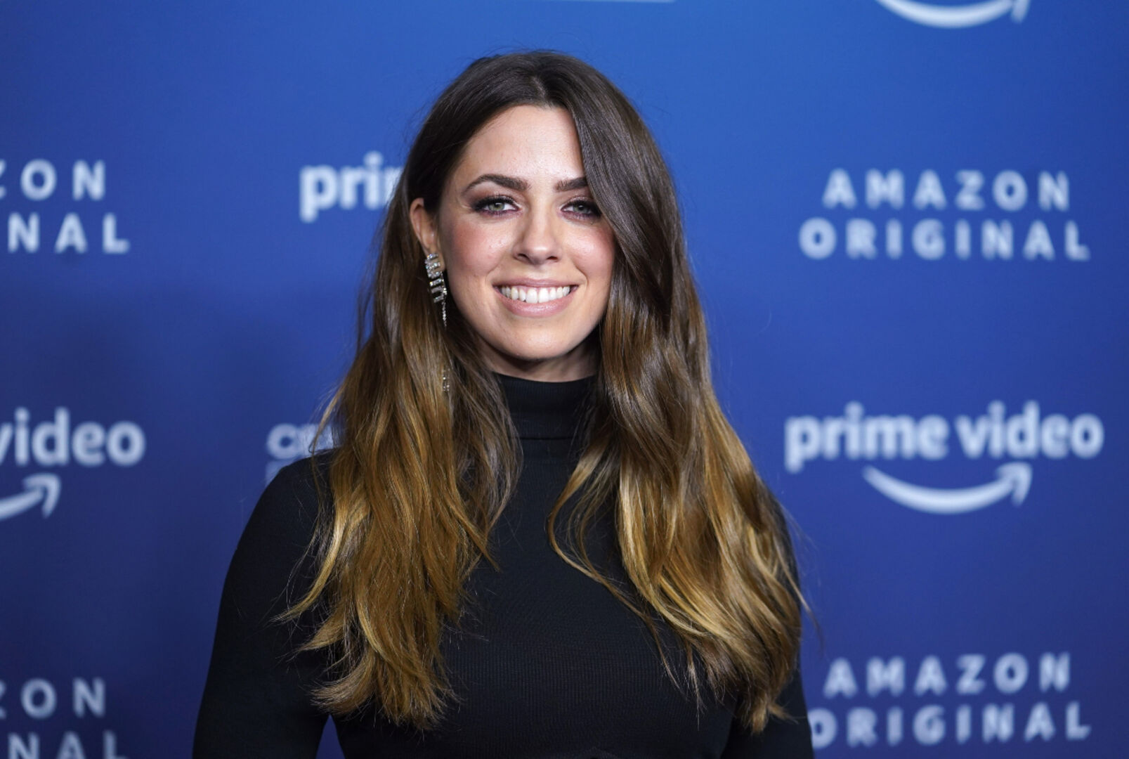 Vanessa Mai bei einer Amazon Prime-Premiere, schwarz gekleidet mit silbernen Ohrringen vor blauem Hintergrundit