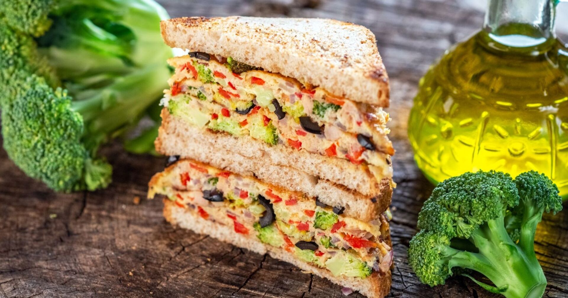 Nahaufnahme eines gestapelten Brokkoli-Sandwiches mit Paprika, Zwiebeln und Oliven, daneben frischer Brokkoli und eine Flasche Olivenöl auf Holztisch.