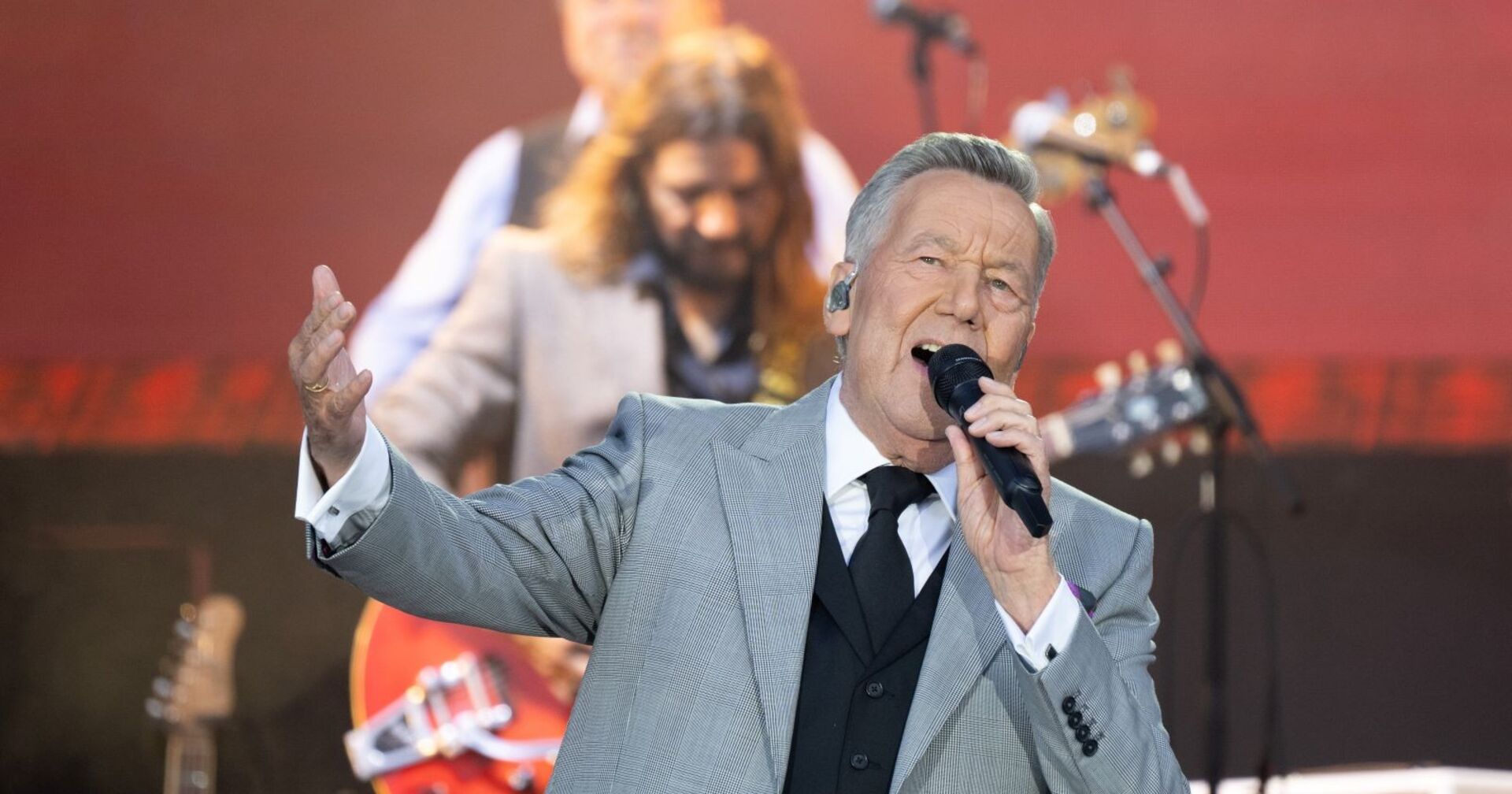 Roland Kaiser singt live auf der Bühne, im grauen Anzug mit Mikrofon in der Hand, Musiker im Hintergrund.