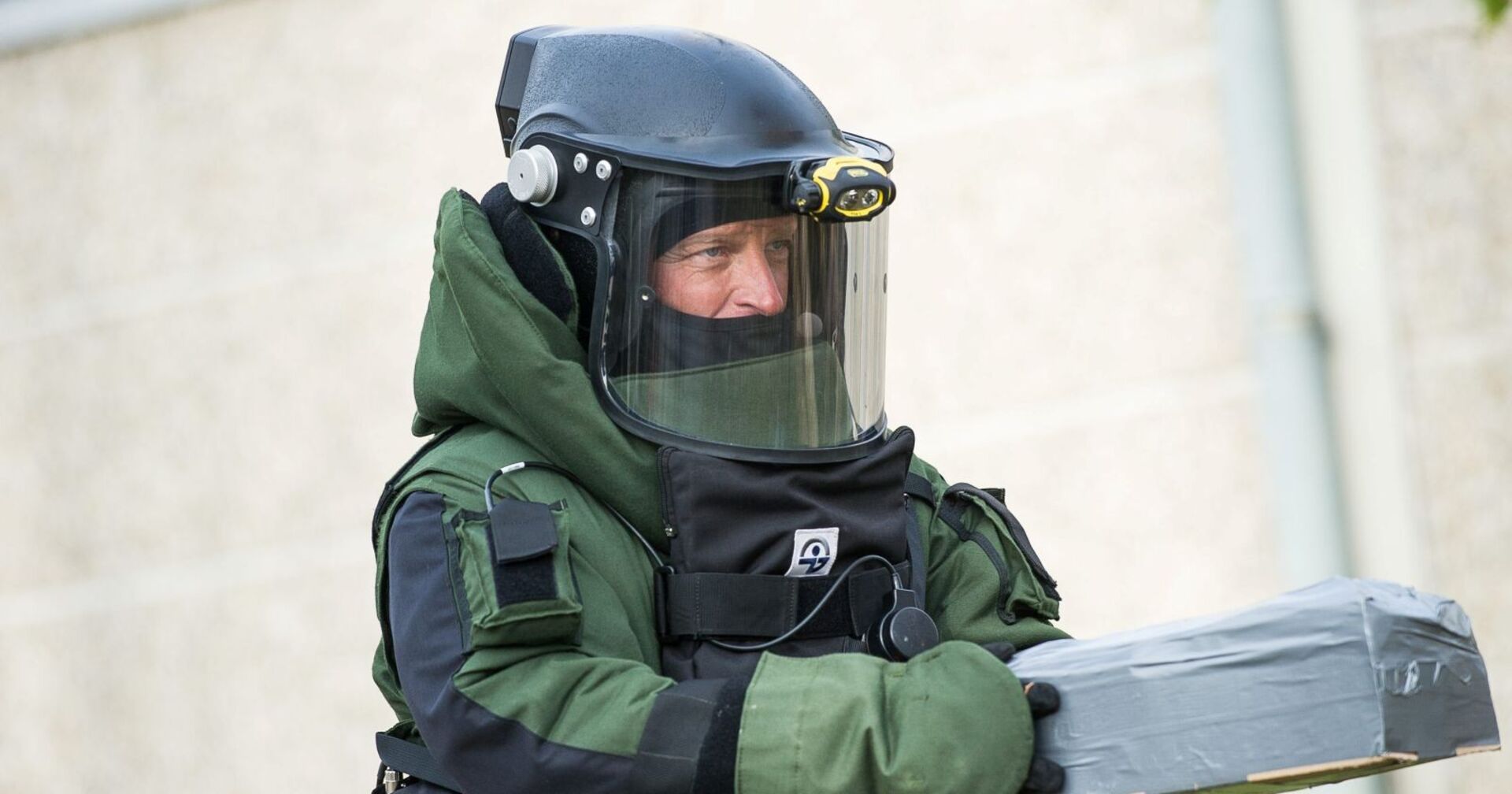 Sprengstoffexperte in schwerer Schutzausrüstung mit Helm und Visier hält ein gesichertes, mit Klebeband umwickeltes Objekt in den Händen.