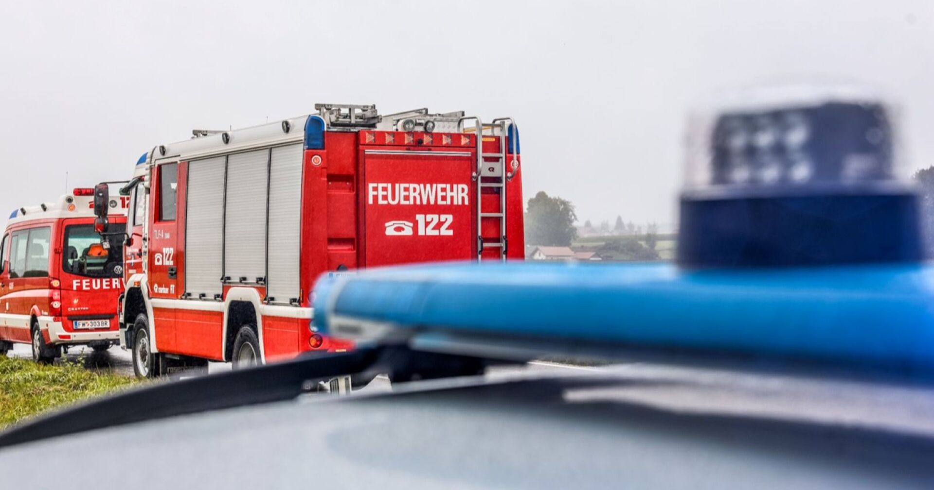 Zwei rote Feuerwehrfahrzeuge mit der Aufschrift „FEUERWEHR 122“ stehen im Einsatz auf einer Landstraße, im Vordergrund ist verschwommen ein blaues Blaulicht eines Polizeifahrzeugs zu sehen.
