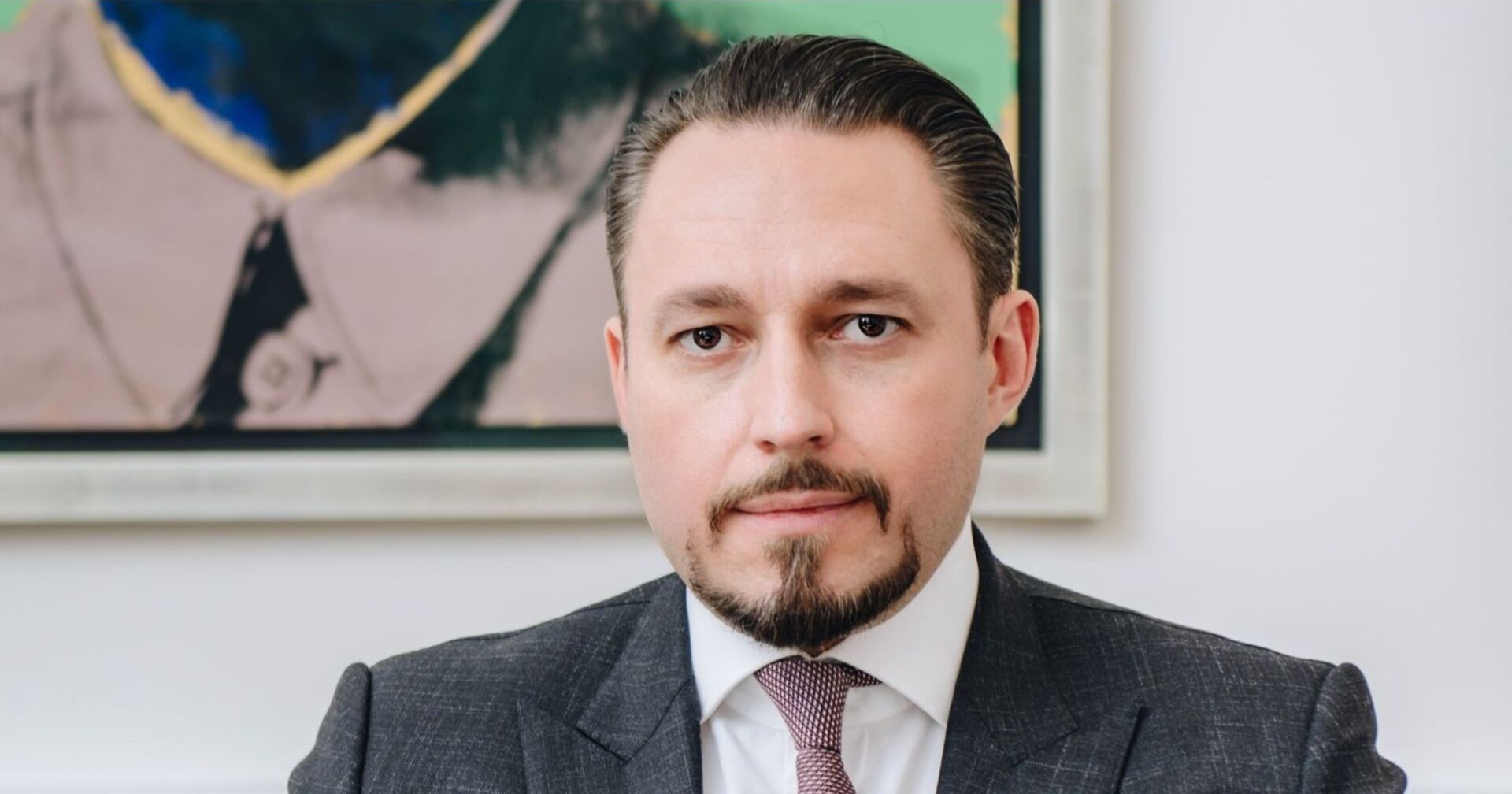 Ein Mann in Anzug und Krawatte blickt direkt in die Kamera. Er hat streng zurückgekämmtes Haar, einen gepflegten Bart und ernsten Gesichtsausdruck. Im Hintergrund ist ein modernes Gemälde zu sehen.