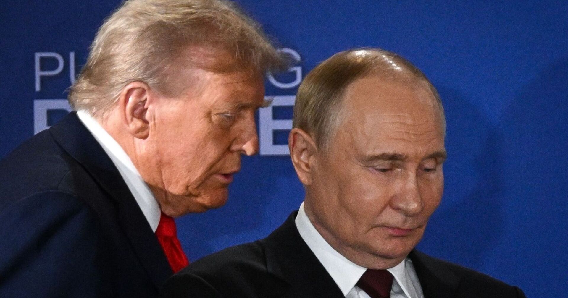 Donald Trump spricht dicht neben Wladimir Putin, der mit ernster Miene nach unten blickt. Beide tragen Anzug, im Hintergrund eine blaue Wand mit dem Schriftzug "Pursuing Peace".