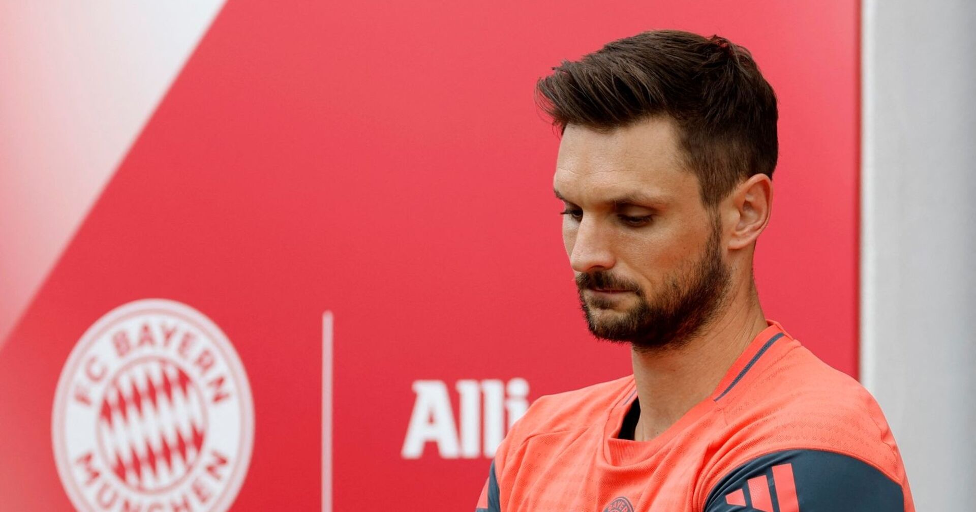 Torwart Sven Ulreich im Trainingsdress des FC Bayern, mit gesenktem Blick vor rotem Hintergrund mit Vereinslogo.