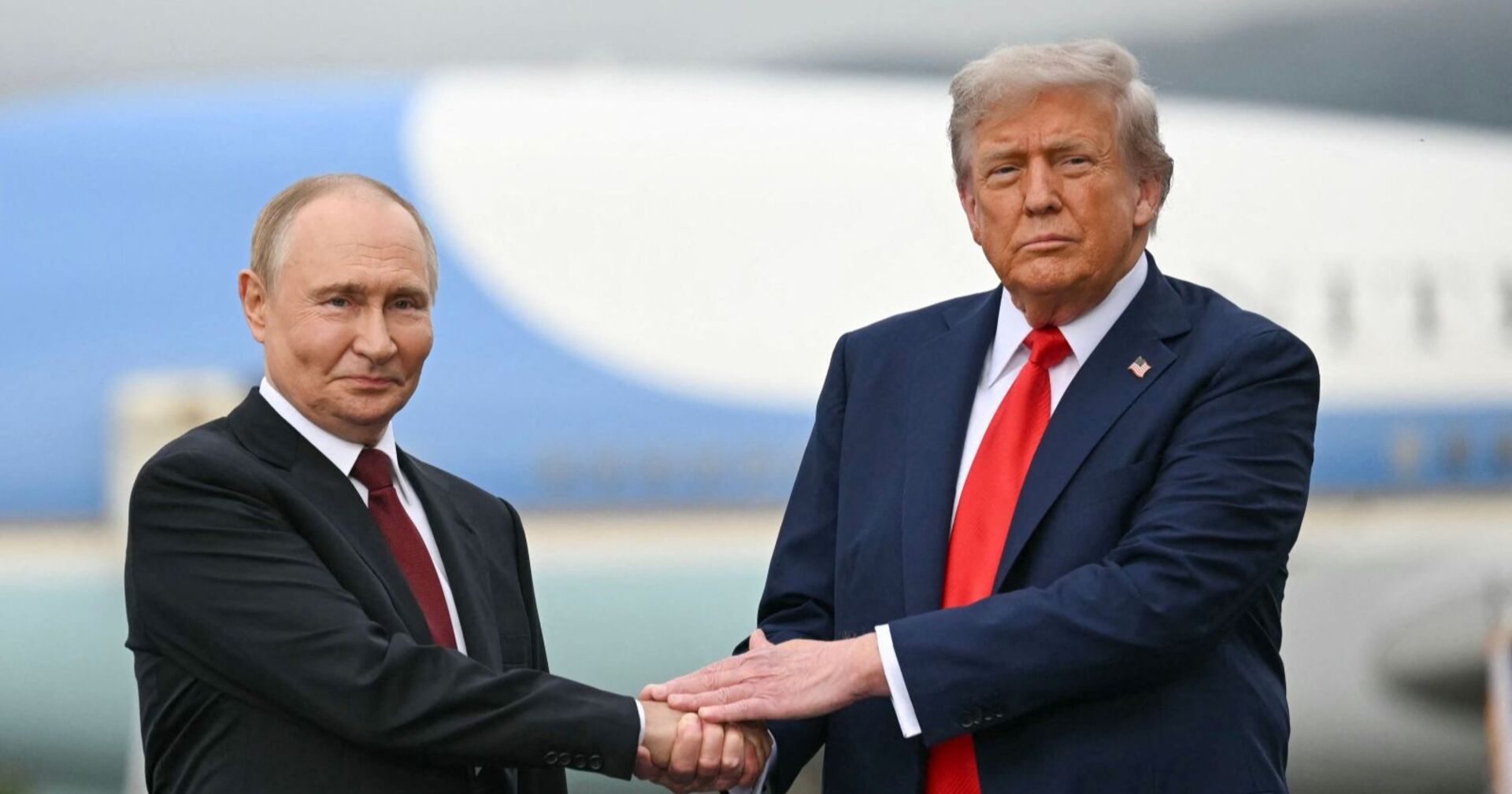 Donald Trump und Wladimir Putin stehen einander gegenüber und schütteln sich vor einem Flugzeughintergrund die Hand. Beide tragen dunkle Anzüge mit roter Krawatte.