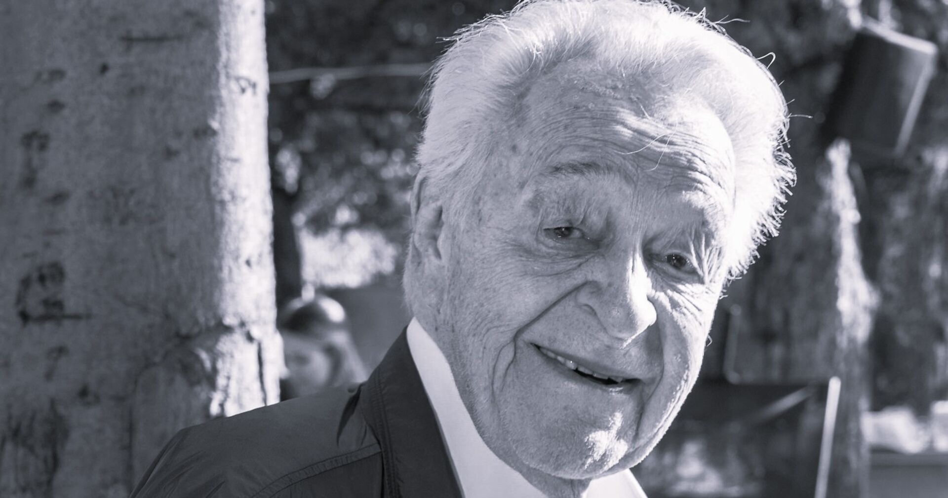 Harald Serafin mit weißem Haar lächelt freundlich in die Kamera; er trägt ein Jackett und steht im Freien neben einem Baum.