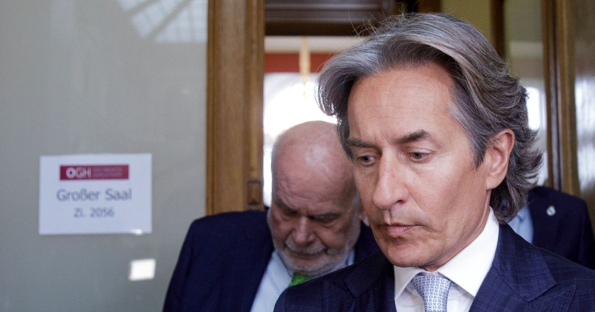 Karl-Heinz Grasser in Anzug und Krawatte verlässt ein Gerichtsgebäude, im Hintergrund ein Schild mit der Aufschrift „OGH – Großer Saal“.