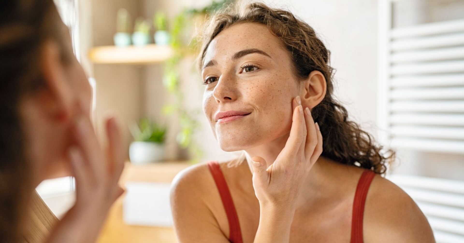 Eine Frau mit Sommersprossen, langen braunen Haaren sieht in den Spiegel. Sie trägt ein rotes Leibchen. Im Hintergrund sieht man Kosmetikarikel.