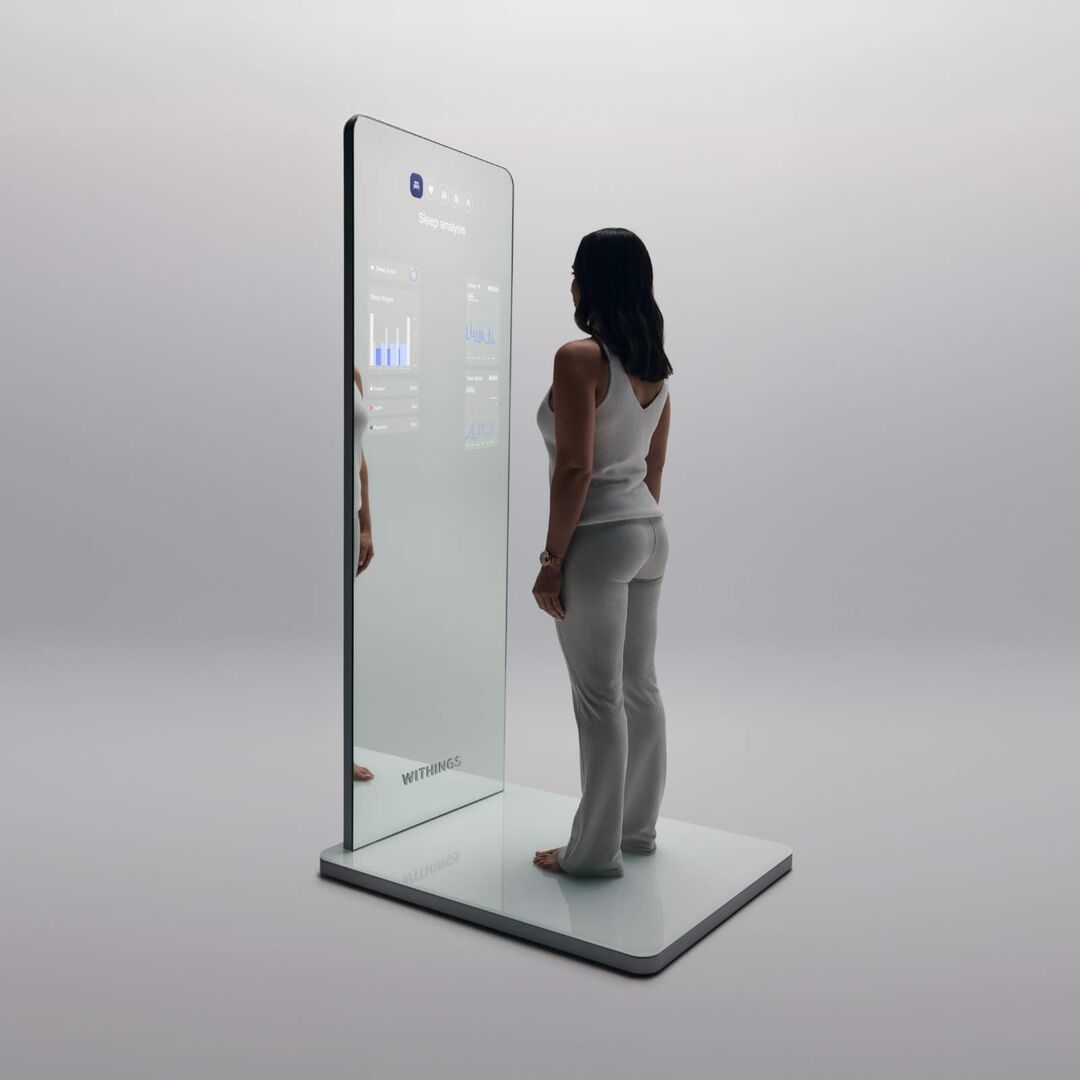 Eine Frau steht barfuß vor einem modernen, bodenstehenden Smart-Spiegel der Marke Withings, der Gesundheitsdaten wie Schlafanalyse auf dem Display anzeigt.