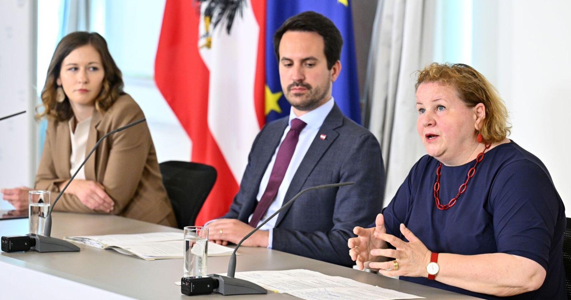 Drei Personen sitzen bei einer Pressekonferenz an einem Tisch mit Mikrofonen und Unterlagen, im Hintergrund sind österreichische und EU-Flaggen zu sehen.