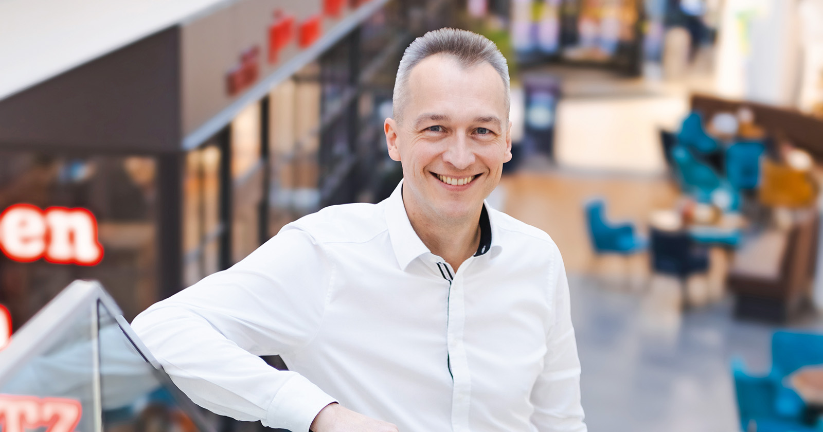 Ein Mann in weißem Hemd lächelt in die Kamera, steht in einem modernen Einkaufszentrum mit Geschäften und Sitzbereichen im Hintergrund.
