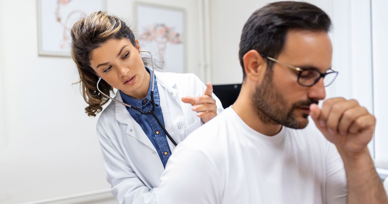 Eine junge Ärztin mit langen, braunen Haaren untersucht mit einem Stethoskop einen Mann. Der Mann trägt eine Brille, hat einen Vollbarte und hustet in seine Faust.