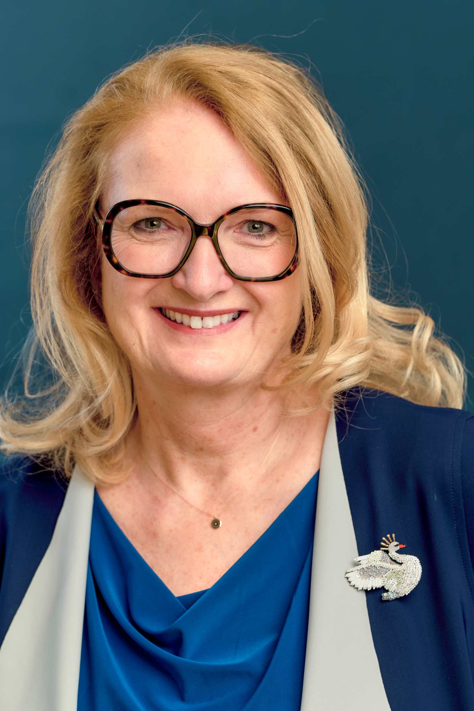 Porträt einer lächelnden Frau mit blonden Haaren und Brille vor blauem Hintergrund. Sie trägt ein blaues Oberteil, ein dunkelblaues Jackett mit hellgrauem Revers und eine Brosche in Form eines silbernen Vogels.
