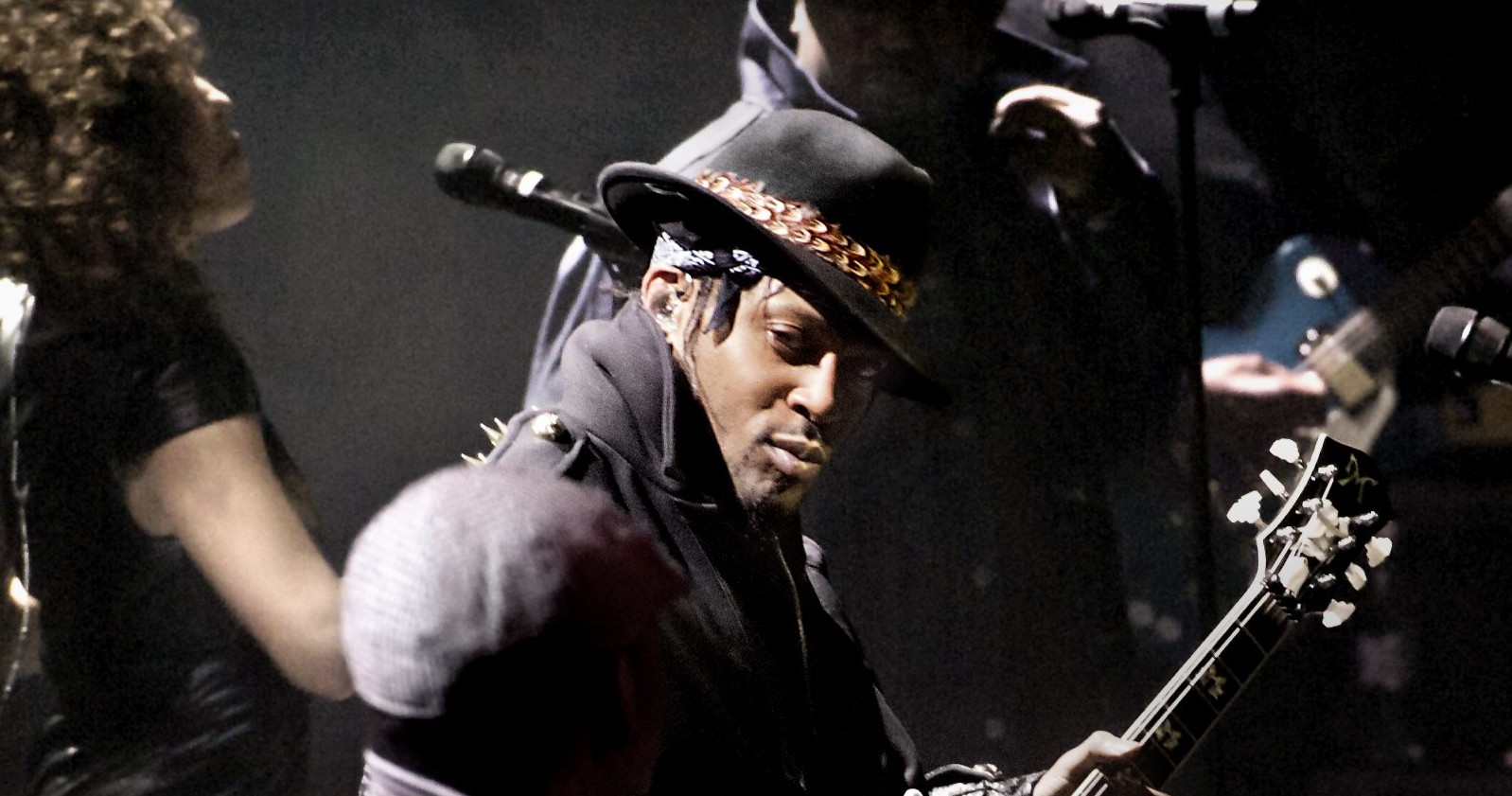 D’Angelo steht auf der Bühne, trägt einen schwarzen Hut mit goldener Kette und spielt Gitarre, während Bandmitglieder im Hintergrund zu sehen sind.