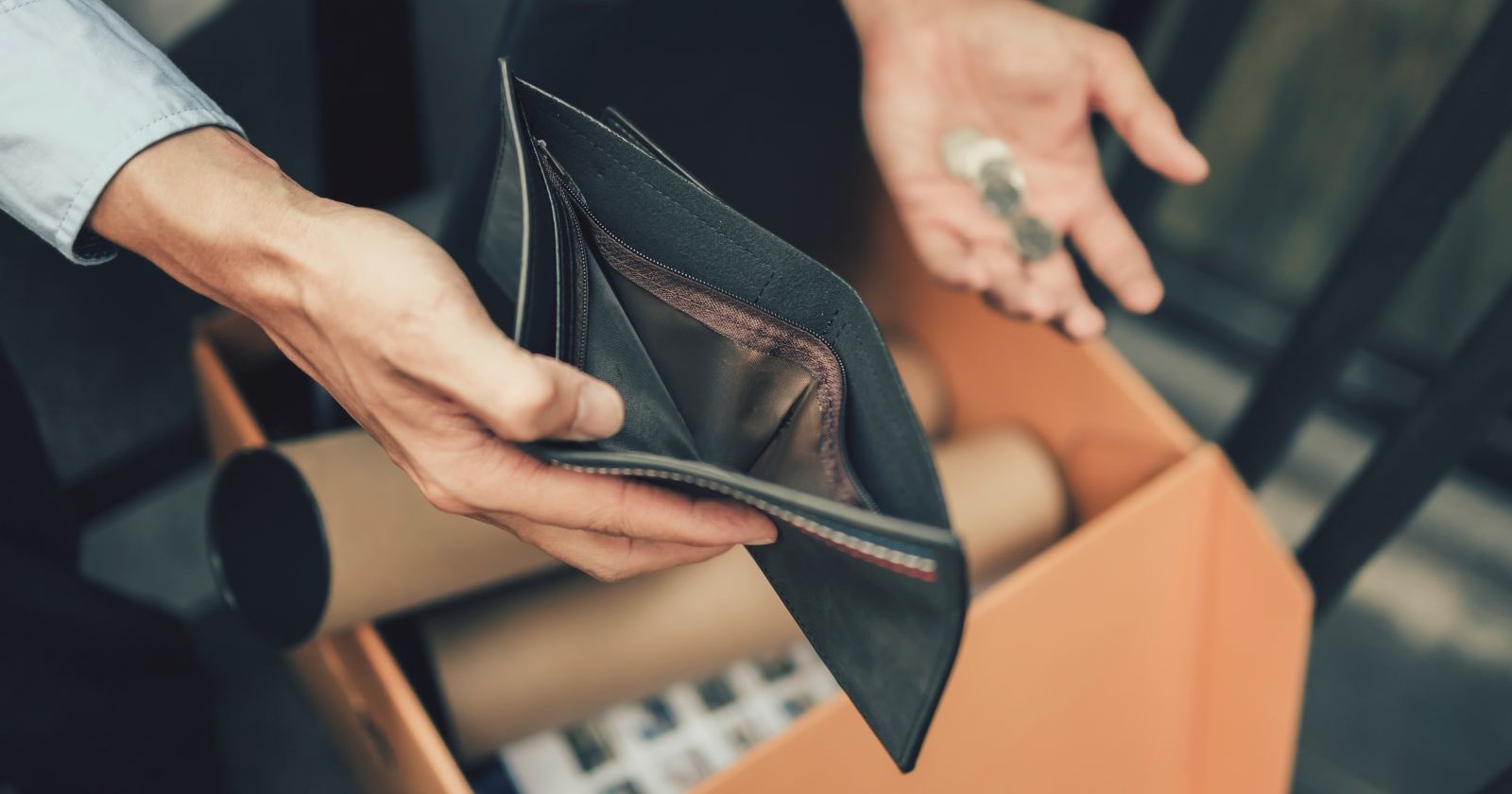 Eine Hand hält eine leere Geldasche. In der anderen Hand sind einige wenige Münzen zu sehen. Unter den Händen ist ein Karton zu sehen, der mit Büroartikeln gefüllt ist.