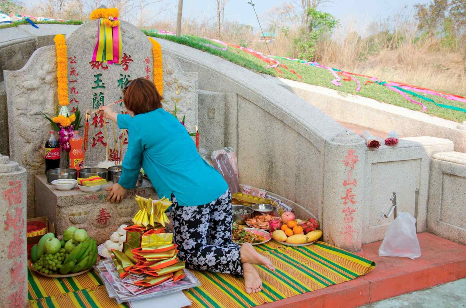 Frau kniet vor einem chinesischen Grabstein mit Opfergaben, Früchten und Räucherstäbchen während des Qingming-Festes.