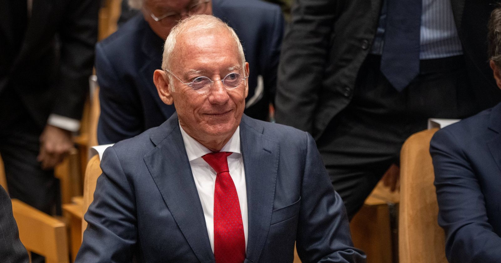 Isak Asic mit Brille, dunkelblauem Anzug und roter Krawatte sitzt zwischen anderen Anzugträgern in einem Konferenz- oder Veranstaltungssaal.