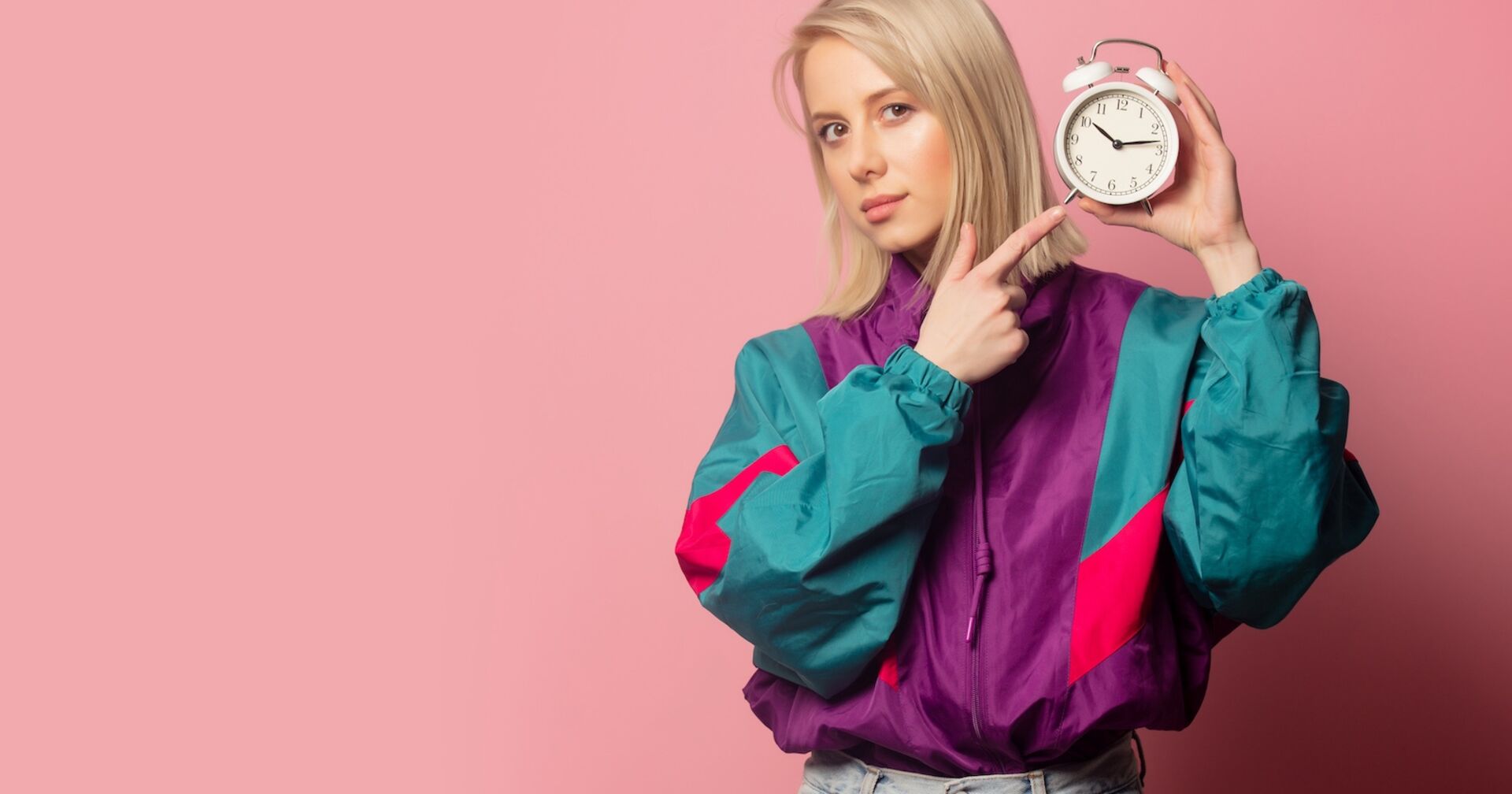 junge blonde Frau in einem bunten Blouson und mit einem klassischen Wecker in der Hand