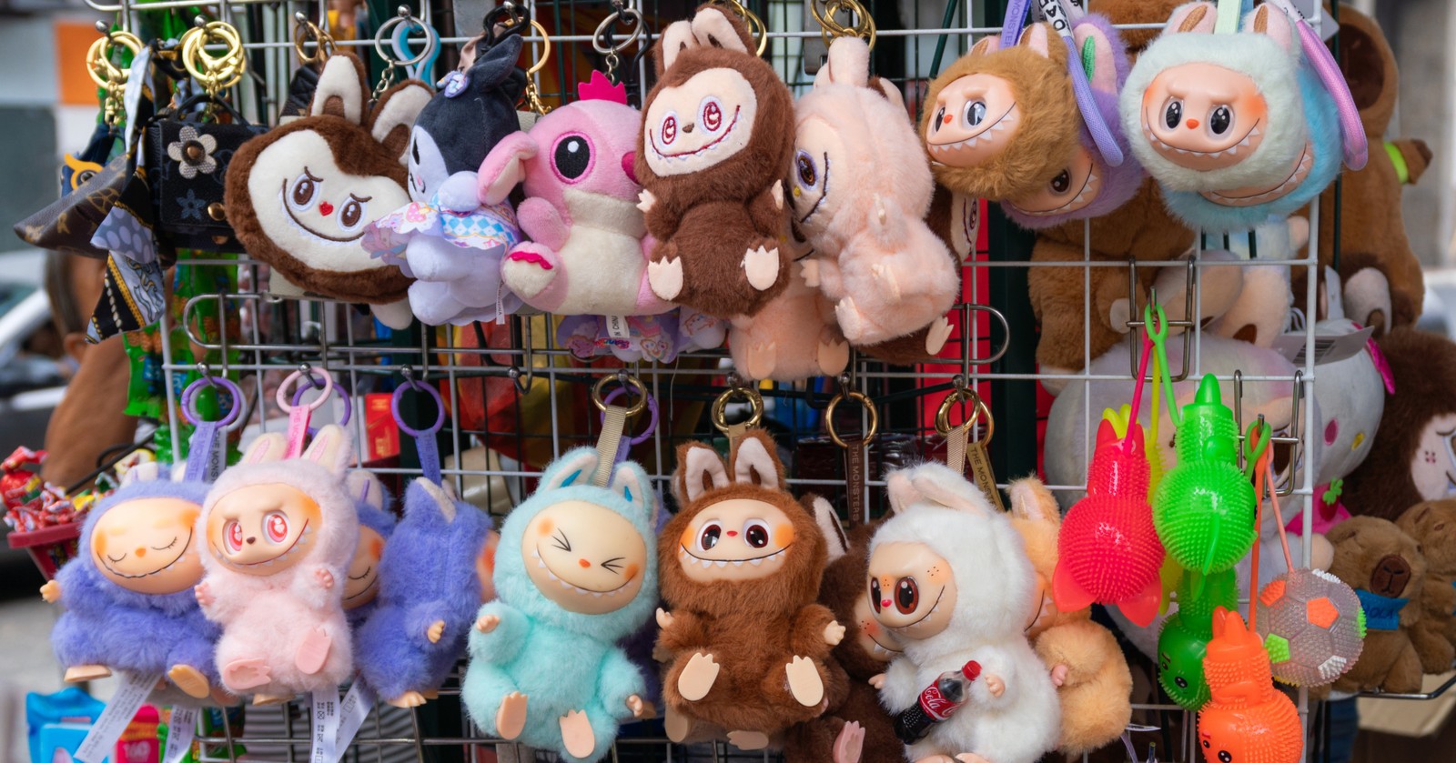 Mehrere bunte Plüschfiguren und Schlüsselanhänger hängen an einem Verkaufsständer auf der Straße, während zwei Personen Geld austauschen.