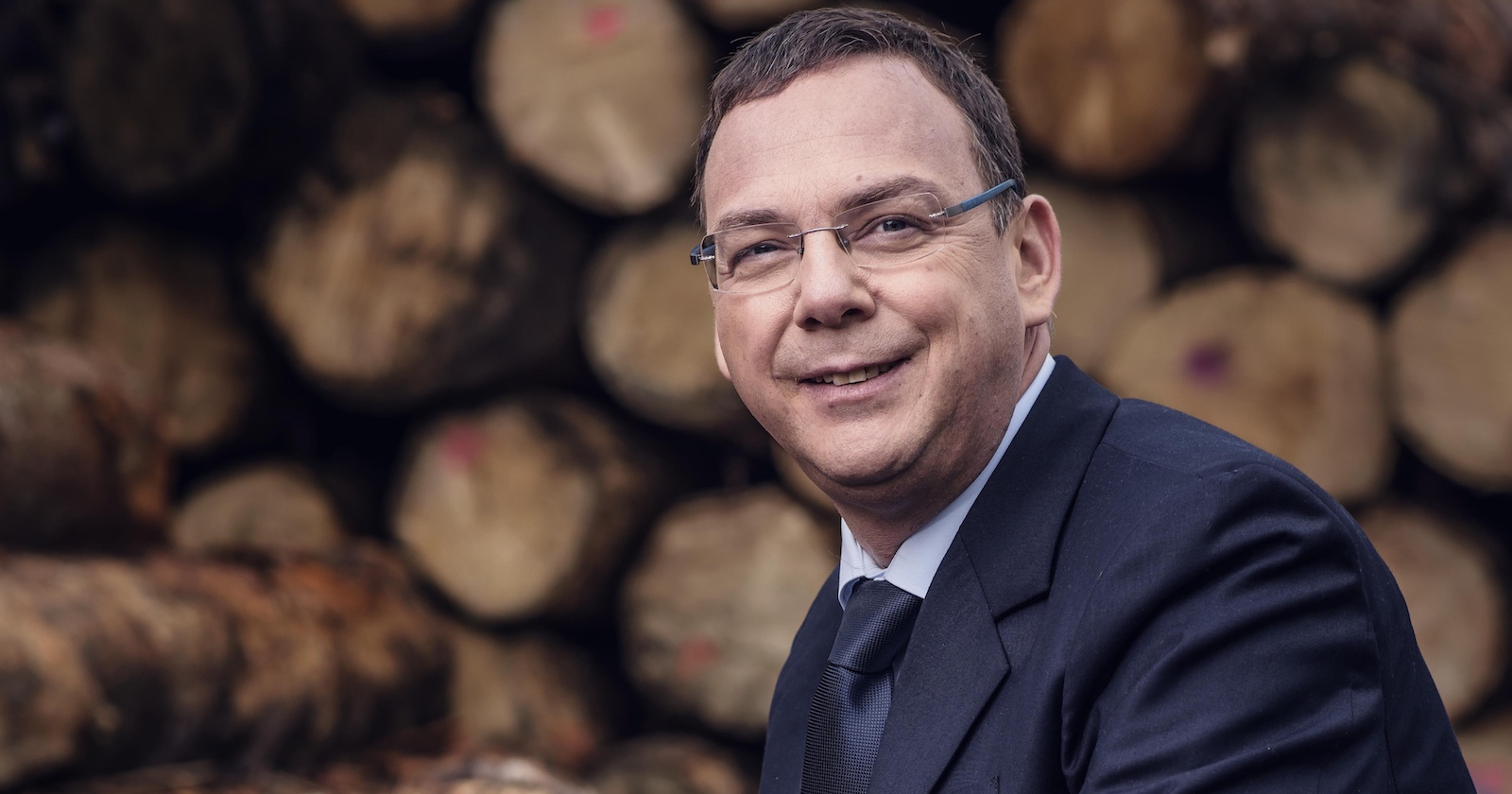 Michael Pfeifer in Anzug und Brille vor einem Holzstoß