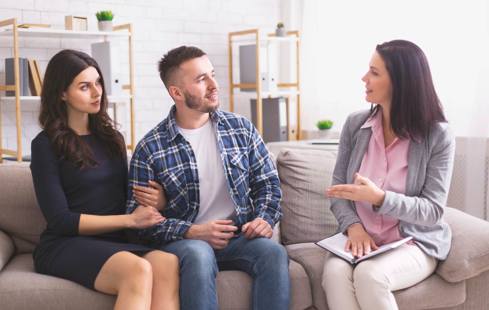 Ein junges Paar sitzt auf einem Sofa und spricht mit einer Therapeutin oder Beraterin. Die Frau hält ein Notizbuch in der Hand und erklärt etwas, während das Paar aufmerksam zuhört.