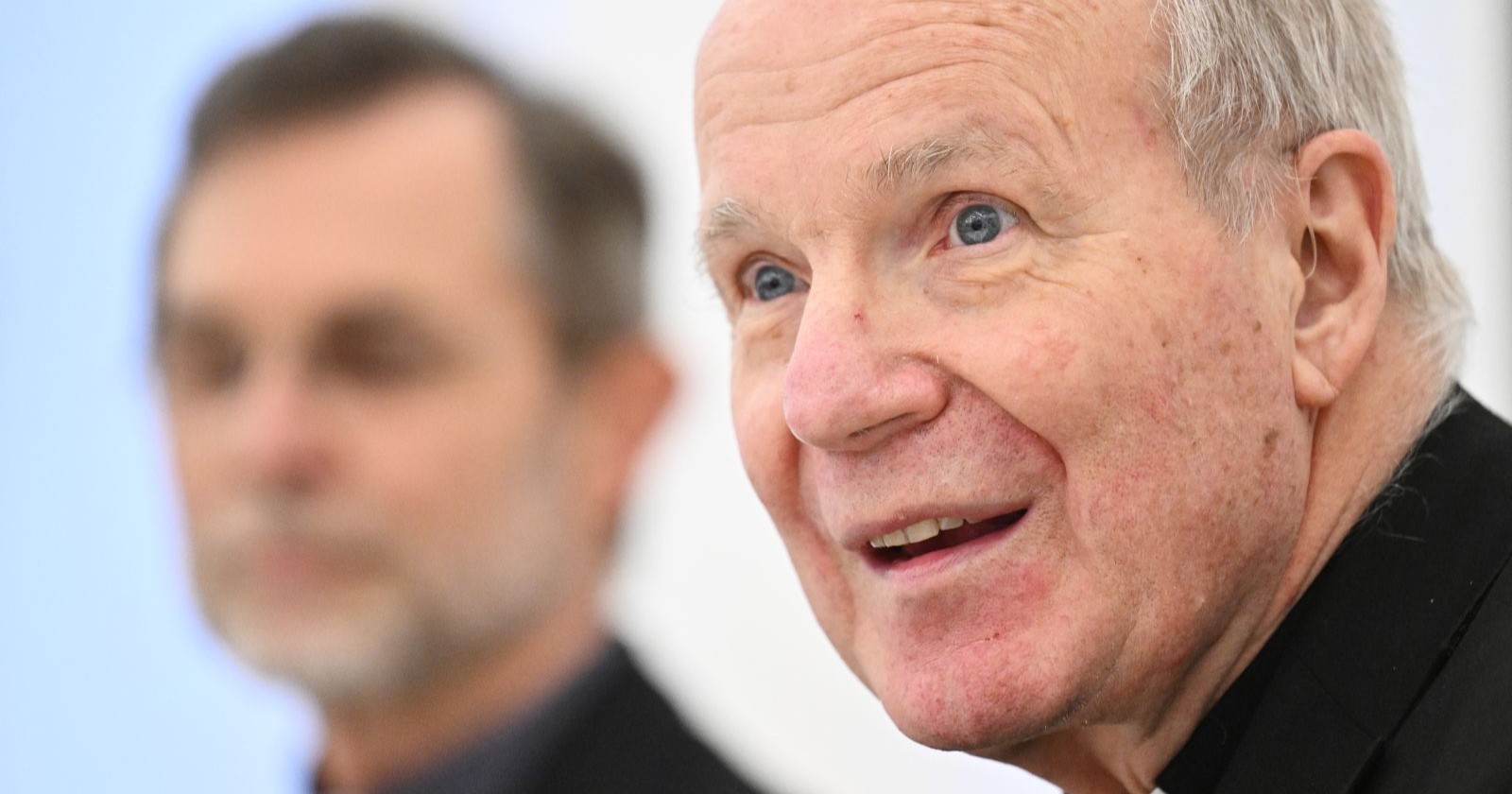 Schönborn im Porträt lächelnd und sprechend bei einer Pressekonferenz zu seiner Nachfolge, im Hintergrund Grünwidl unscharf verschwommen.