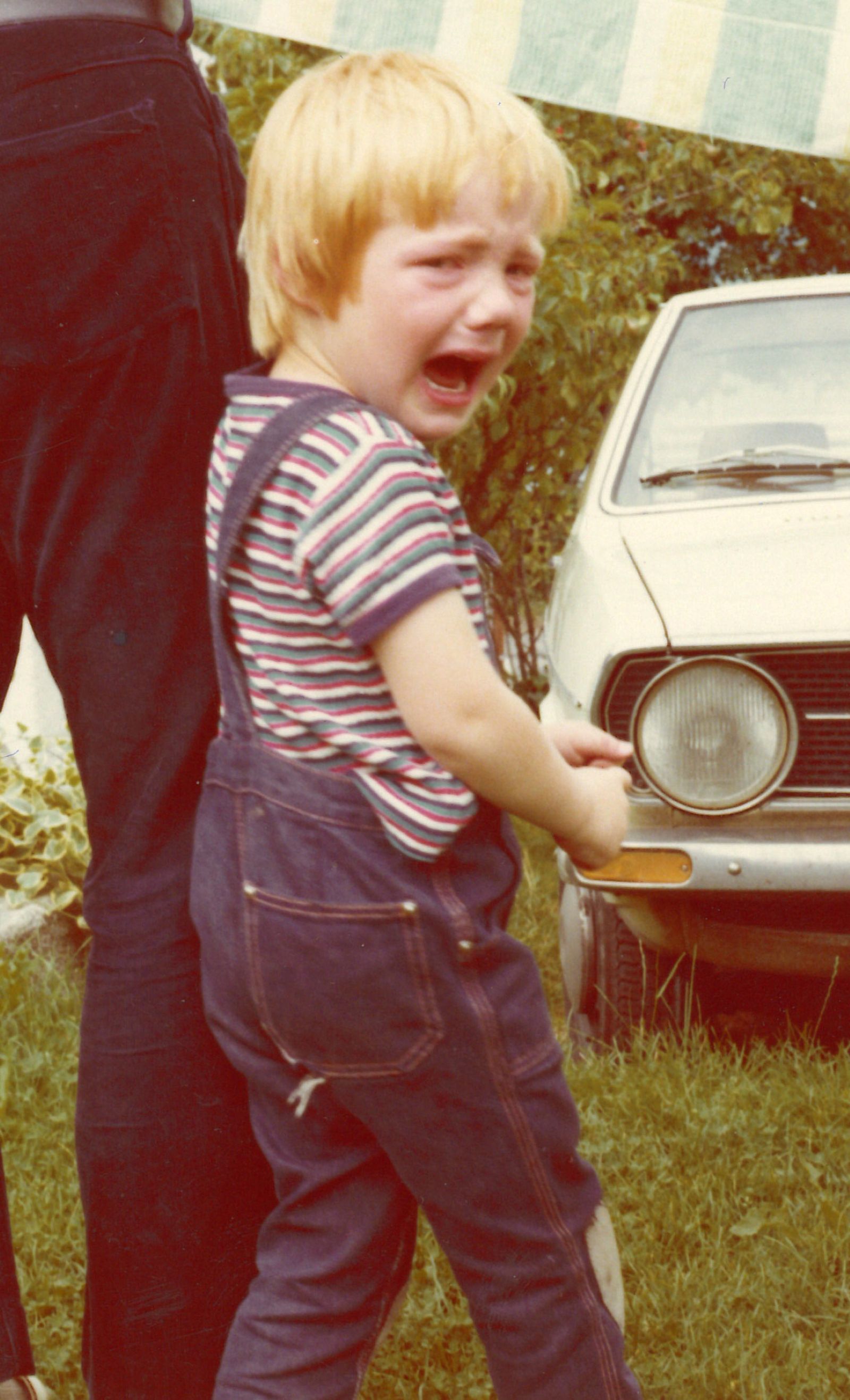 Simon Schwarz als kleiner Bub in Latzhose im Garten, im Hintergrund ist ein Auto einer undefinierbaren Marke zu sehen