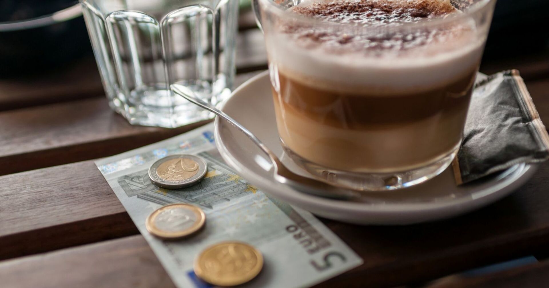 Eine Tasse Kaffee und ein Glas weiter, darunter ein Fünf-Euro-Schein und einige Münzen als Symbol für die Debatte um die Trinkgeld-Debatte.