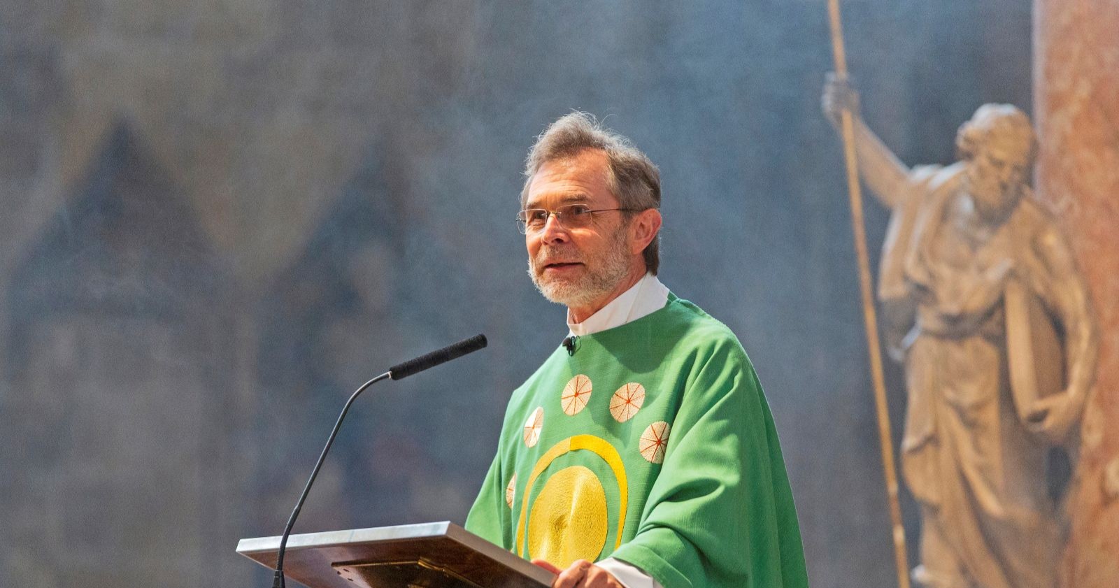 Josef Grünwidl in grüner liturgischer Kleidung predigt am Ambo, hinter ihm steht eine Statue mit Stab.