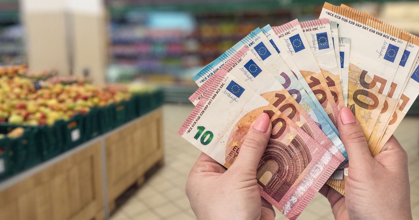 Eine Person hält mehrere Euro-Banknoten in der Hand, im Hintergrund sind Obstregale in einem Supermarkt zu sehen; Themenbild für die neuen EU-Regeln zur Behebung von Bargeld im Supermarkt.