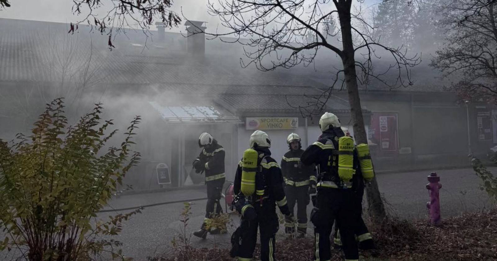 Feuerwehrleute vor dem völlig verrauchten Gebäude der brennenden Tennishalle in Klagenfurt-Welzenegg