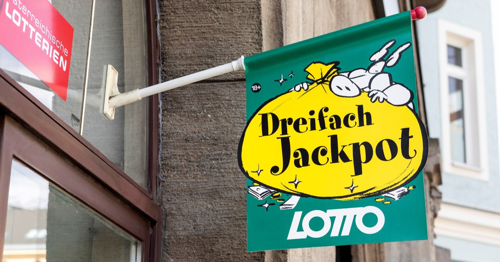 Werbeschild der Österreichischen Lotterien mit der Aufschrift „Dreifach Jackpot“ vor einer Lotto-Annahmestelle.