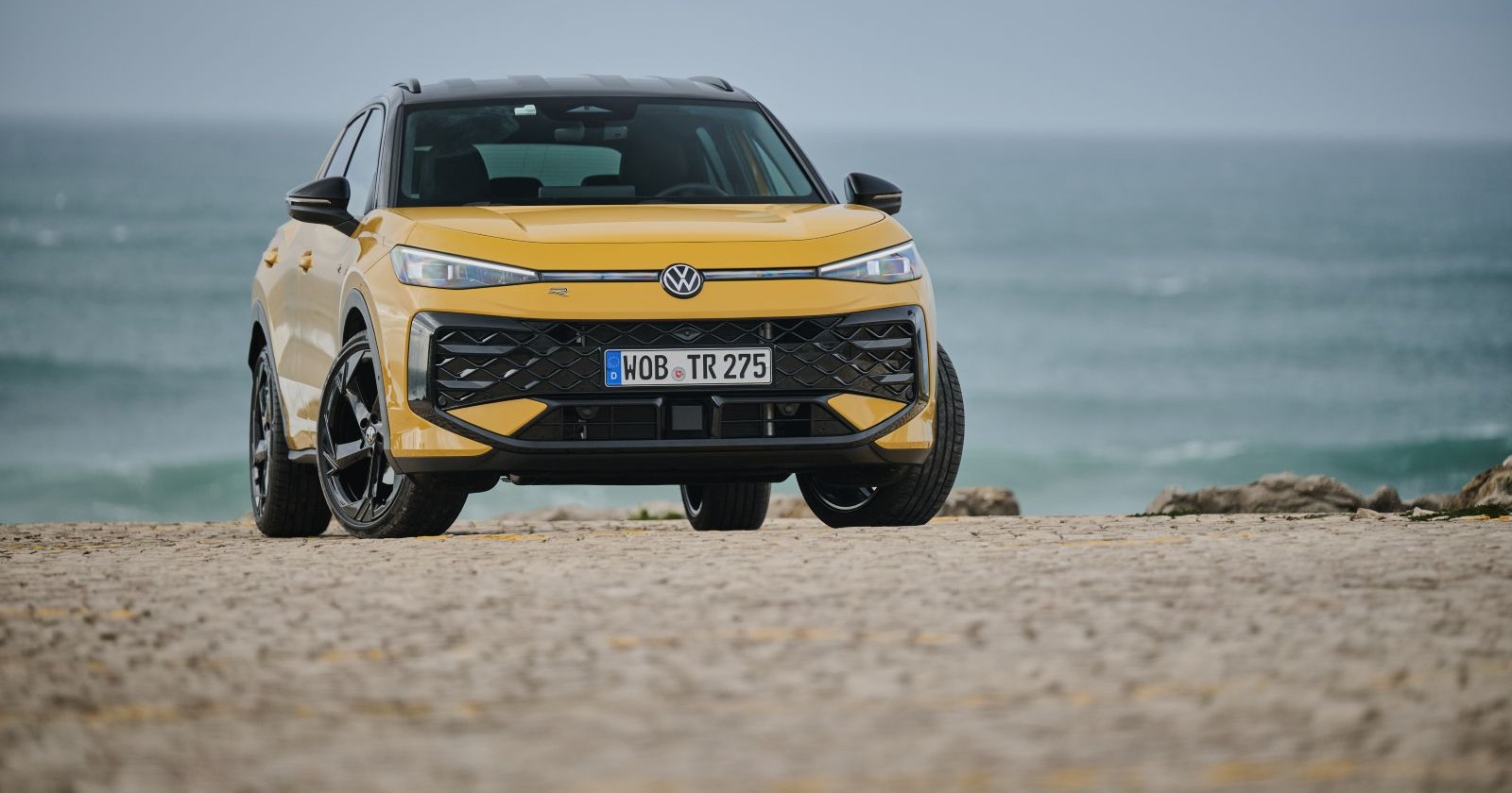 Ein gelber VW T-Roc parkt vor dem Meer, das kaum Wellen aufweist. Der Untergrund auf dem das Auto steht besteht aus Stein.