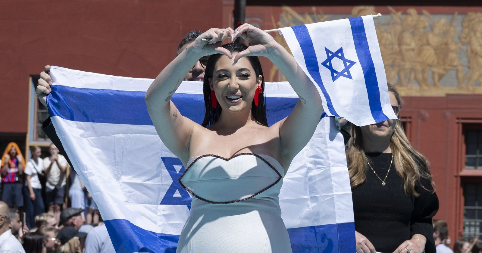 Die israelische ESC-Künstlerin zeigt ein Herz in die Menge, während hinter ihr mehrere Personen israelische Flaggen halten – ein deutliches Zeichen der Unterstützung kurz vor ihrem Auftritt.